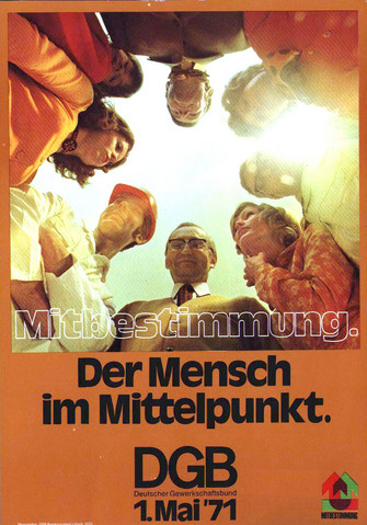 1. Mai 1971: Motiv: 8 Menschen verschiedener Berufsguppen im Kreis sind aus der Froschperspektive zu sehen und die Sonne beleuchtet die Szene von oben. Text unter dem Foto: "Mitbestimmung. Der Mensch im Mittelpunkt. DGB - Deutscher Gewerkschaftsbund - 1. Mai '71"