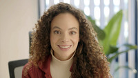 Portraitbild von einer lächelnden Frau mit langen Locken an einem Schreibtisch.