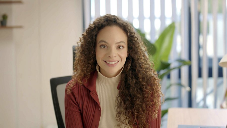 Portraitbild von einer lächelnden Frau mit langen Locken an einem Schreibtisch.