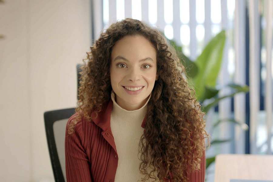 Portraitbild von einer lächelnden Frau mit langen Locken an einem Schreibtisch.