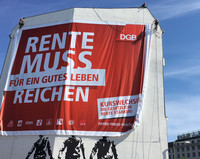 Arbeiter rollen ein Banner mit der Aufschrift "Rente muss für ein gutes Leben reichen" an einer Hausfassade aus. Darunter eine Grafitti mit 3 Männerfiguren