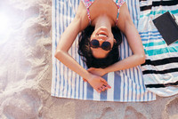 Foto einer Frau mit Sonnenbrille, die am Strand auf einem Badehandtuch liegt und lacht. 