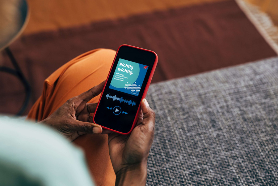 Frau sitzt auf der Couch mit einem Smartphone in der Hand und hat den Podcast richtig.wichtig geöffnet.